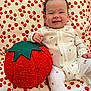 Nora is registered to the contest to win money with this photo: baby, child, onesie, plush_toy, tomato_pillow, red, green, white, pattern, cherry_pattern, blanket, smile, happy, sitting, indoor, cute, face, hand, hair, soft_toy