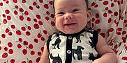 Nora is registered to the contest to win money with this photo: baby, smiling, onesie, dog_pattern, black_and_white, pillow, tomato_print, red, white, infant, happy, cute, indoor, child, headband, bow, sitting, face, skin, arm