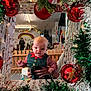 baby, child, holiday_clothing, christmas_decorations, red_ornaments, green_garland, indoor, person, hand, wooden_fence, rainbow_drawing, playroom, festive, window_frame, gingerbread_ornament, holiday_spirit, smiling, festive_outfit, celebration, decor