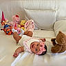 baby, infant, pink_headband, white_outfit, stuffed_animals, teddy_bear, bunny, tiger, party_hat, bed, pillow, smiling, happy, lying_down, soft_toys, indoor, child, cute, playful, cozy
