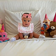 Valentina is registered to the contest to win money with this photo: baby, bib, bow, celebration, child, cute, home, indoor, infant, octopus_toy, party_hats, pink_headband, portrait, sitting, smiling, soft_toys, stuffed_animals, teddy_bear, tigger_toy, white_bed