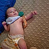 baby, infant, child, lying_down, couch, blanket, bib, pacifier, diaper, wristband, blue_clothing, patterned_fabric, indoor, portrait, face, eyes_open, extended_arm, comfortable, home, safe