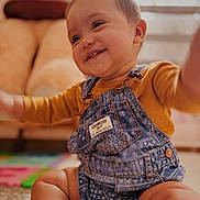 Velincia joined the competition — help win amazing prizes! baby, child, clothing, cozy, cute, denim_overalls, face, happy, home, indoors, infant, legs, mustard_shirt, person, portrait, rug, sitting, smiling, soft_light, young_child