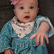 Violet is registered to the contest to win money with this photo: baby, child, infant, face, hand, headband, flower, dress, bib, pattern, bird_pattern, blue_clothing, pink_accessory, curious_expression, sitting, portrait, indoor, cozy, cute, small_hand