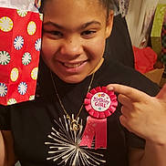 Sophia joined the competition — help win amazing prizes! child, birthday, smiling, gift_bag, ribbon, hair_bun, black_tshirt, necklace, indoor, celebration, happy, person, portrait, clothing, jewelry, hand, chair, purple_pants, decor, party