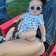 Walker is registered to the contest to win money with this photo: adult, baby, casual_clothing, chair, child, footwear, grass, greenery, hand, infant, legs, onesie, outdoor, person, red_chair, relaxing, sandal, summer, sunglasses, sunny