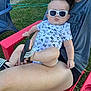 adult, baby, casual_clothing, chair, child, footwear, grass, greenery, hand, infant, legs, onesie, outdoor, person, red_chair, relaxing, sandal, summer, sunglasses, sunny