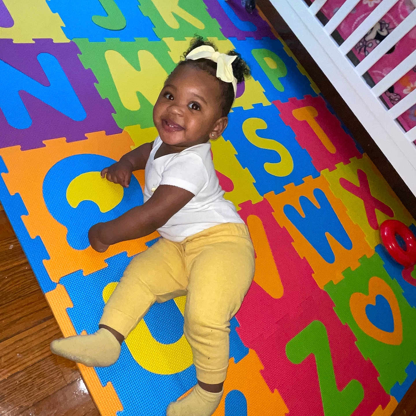 Daliya is registered to the contest to win money with this photo: alphabet_mat, baby, child, colorful, crib, cute, flooring, foam_mat, happy, indoor, person, playroom, smiling, socks, toddler, toy, white_shirt, wooden_floor, yellow_bow, yellow_pants