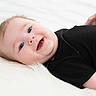 baby, infant, smiling, lying_down, black_clothing, soft_texture, white_background, cute, happy, child, portrait, face, blue_eyes, short_hair, chubby_cheeks, innocence, comfort, relaxation, indoor, closeup