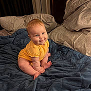 Ayda is registered to the contest to win money with this photo: baby, child, yellow_onesie, blue_blanket, bed, pillow, smile, cute, indoors, soft_lighting, chubby_cheeks, sitting, infant, warm_colors, cozy, happy, portrait, person, furniture, blanket