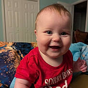 Ayda is registered to the contest to win money with this photo: baby, child, smiling, red_shirt, indoor, bedspread, blue_wall, doorway, closet, happy, portrait, cute, chubby, infant, person, sitting, room, expression, casual, home