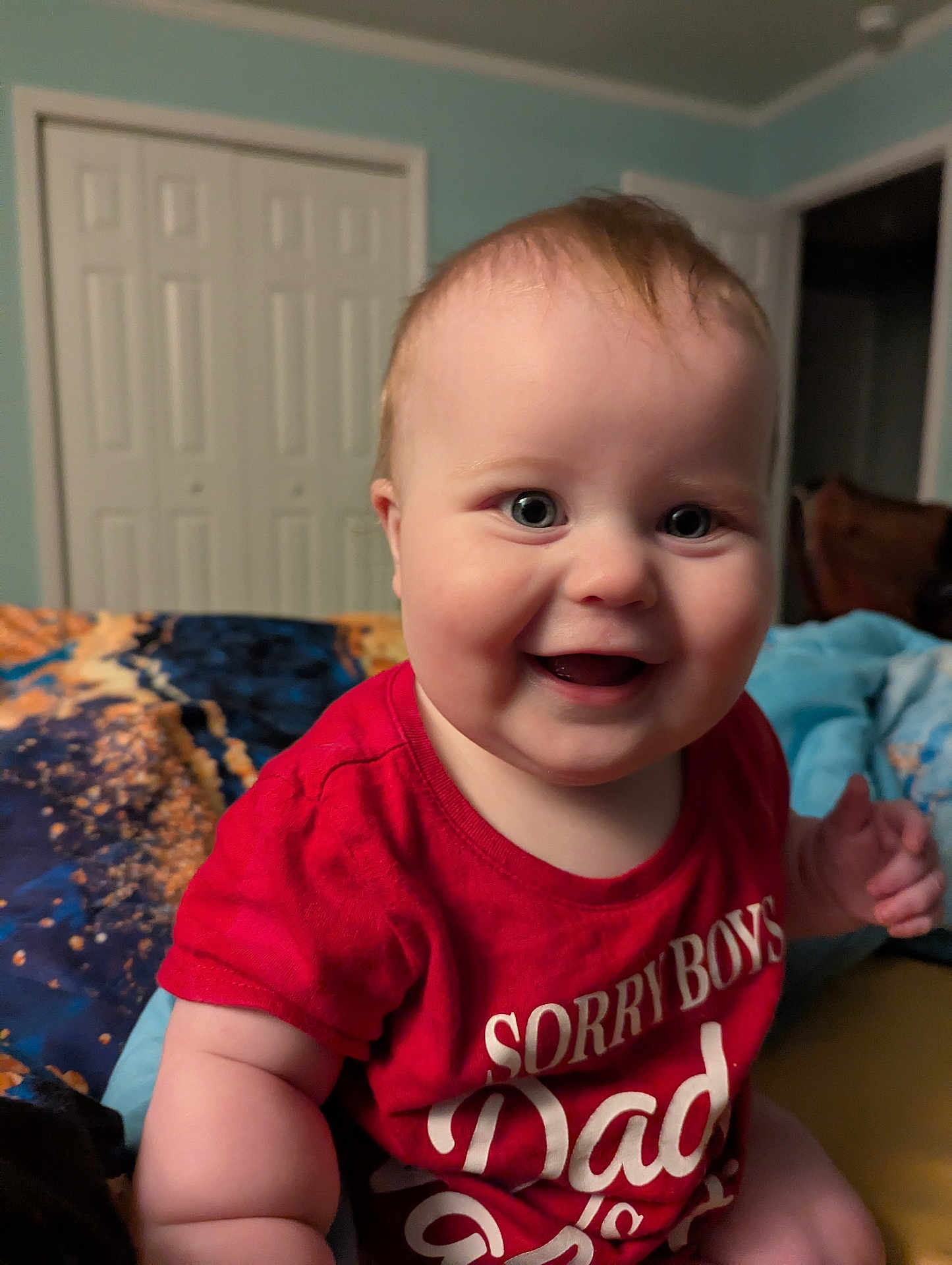 Ayda is registered to the contest to win money with this photo: baby, child, smiling, red_shirt, indoor, bedspread, blue_wall, doorway, closet, happy, portrait, cute, chubby, infant, person, sitting, room, expression, casual, home