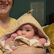 Ayda is registered to the contest to win money with this photo: baby, adult, person, towel, hooded_towel, smile, face, hand, arm, bedroom, door, blanket, yellow_shirt, indoor, curious, holding, comfort, skin, blue_eyes, home