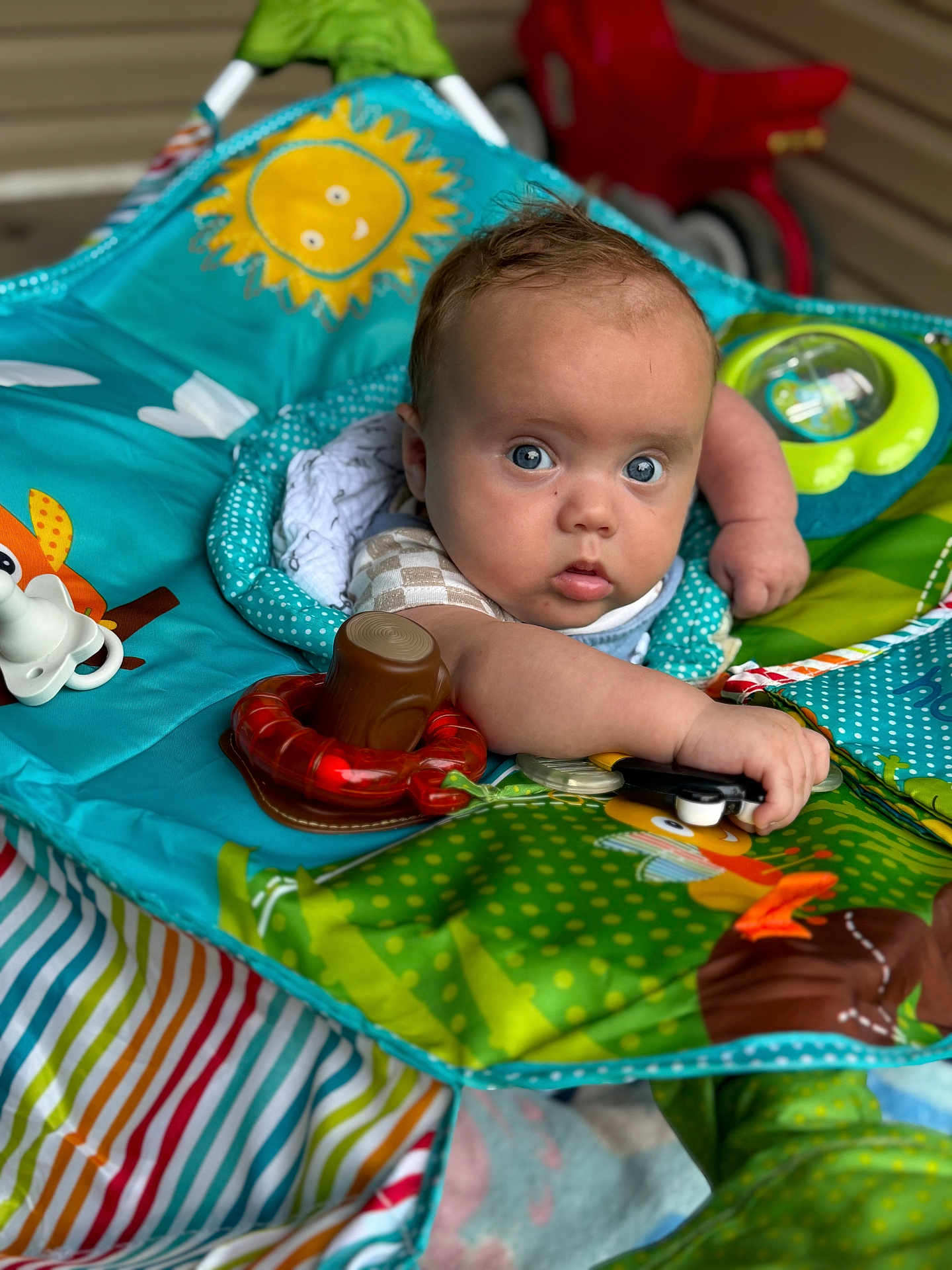 Elias is registered to the contest to win money with this photo: baby, infant, play_mat, toy, colorful, blue_eyes, curious, child, indoor, striped_blanket, cute, small_arm, patterned_clothing, soft_toys, red_toy_car, bright_colors, face, expression, young, playtime