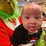 baby, child, face, tongue, costume, grinch, mask, green_hair, red_clothing, holiday, christmas, indoor, person, smile, celebration, festive, holding, background, ceiling_fan, table