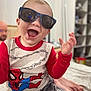 toddler, child, sunglasses, laughing, spiderman_shirt, red_sleeves, gray_pants, indoor, bedroom, happy, smiling, person, casual_clothing, short_hair, playful, cute, fun, young_child, seated, home