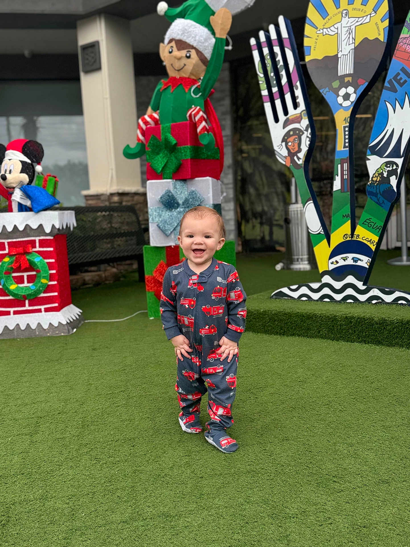 Oliver is registered to the contest to win money with this photo: toddler, child, pajamas, fire_truck, smiling, grass, artificial_grass, holiday_decor, elf, gift_boxes, decorations, outdoor, bench, toy, colorful, festive, playful, happy, person, portrait