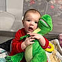 Oliver is registered to the contest to win money with this photo: baby, child, stuffed_toy, dinosaur, plush, red_clothing, indoor, playpen, quilt, toy, cuddling, cute, smiling, person, infant, soft_toy, floor, hand, face, happy