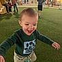 toddler, child, running, green_sweater, beige_shorts, outdoor, artificial_turf, nighttime, group_of_people, shopping_area, happy, smiling, playful, casual_clothing, background_people, storefront, lighting, fun, motion, joy