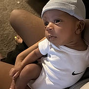 Jah Mauri is registered to the contest to win money with this photo: baby, infant, child, portrait, white_hat, white_clothing, nike_logo, socks, sitting, held, adult, arms, indoors, home, carpet, furniture, close_up, looking_away, calm, photography