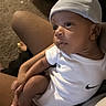 baby, infant, child, portrait, white_hat, white_clothing, nike_logo, socks, sitting, held, adult, arms, indoors, home, carpet, furniture, close_up, looking_away, calm, photography