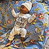baby, infant, child, portrait, lying_down, blanket, blue_blanket, mickey_mouse, cartoon, white_outfit, white_hat, clothing, bed, indoor, face, hands, feet, cute, text_on_clothing, cozy
