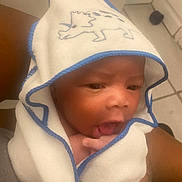 Jarez is registered to the contest to win money with this photo: baby, towel, hooded_towel, dinosaur, infant, face, tongue, hand, person, arm, skin, expression, indoor, floor, tile, holding, cute, child, newborn, soft