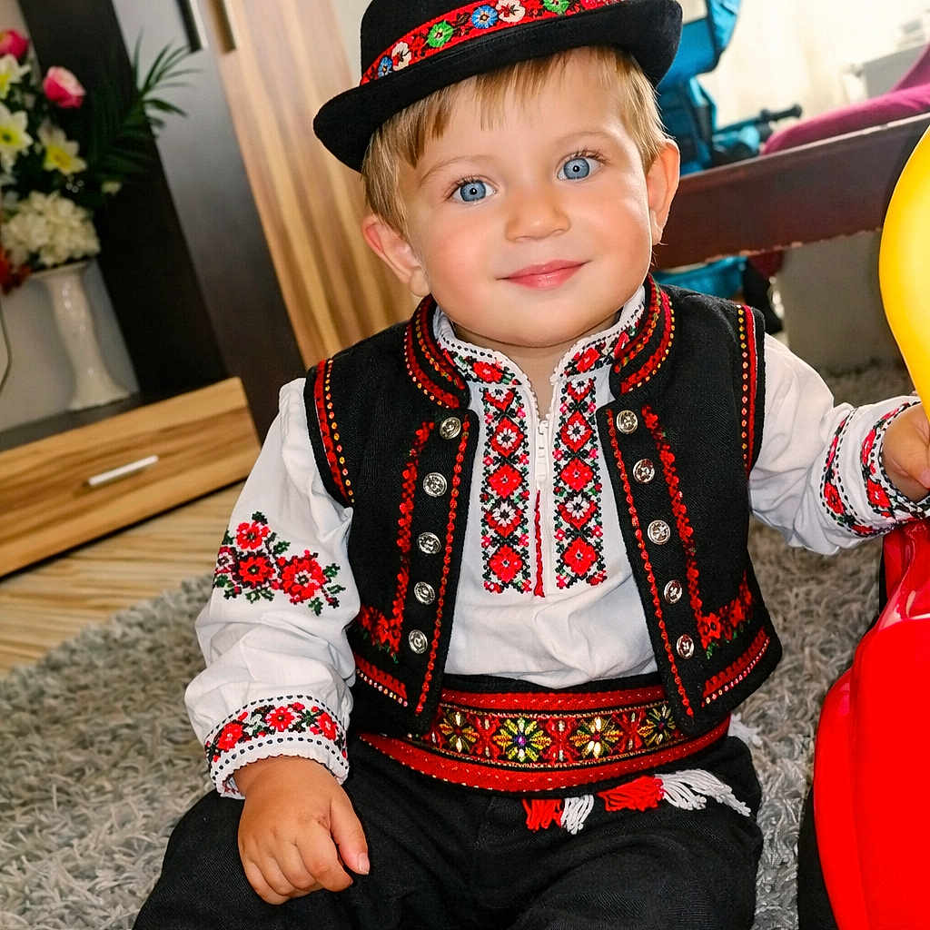 Samuel is registered to the contest to win money with this photo: blue_eyes, boots, cabinet, carpet, child, decor, embroidery, feather, flowers, folk_costume, furniture, hat, indoor, portrait, smiling, toddler, toy_vehicle, traditional_clothing, vest, white_shirt