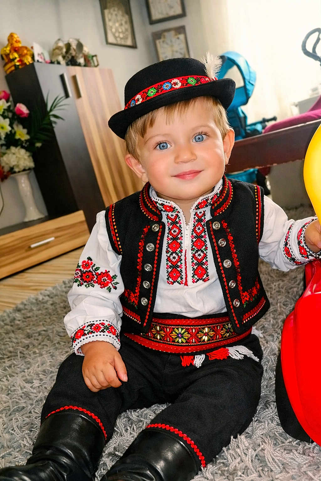 Samuel is registered to the contest to win money with this photo: toddler, child, traditional_clothing, folk_costume, embroidery, hat, feather, blue_eyes, smiling, indoor, carpet, toy_vehicle, boots, vest, white_shirt, decor, flowers, cabinet, furniture, portrait