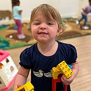 Kimber is registered to the contest to win money with this photo: child, toddler, toy, building_blocks, indoor, playroom, smile, person, short_hair, blue_shirt, young, playing, fun, happy, colorful, floor, background, other_children, plastic_toys, casual_clothing