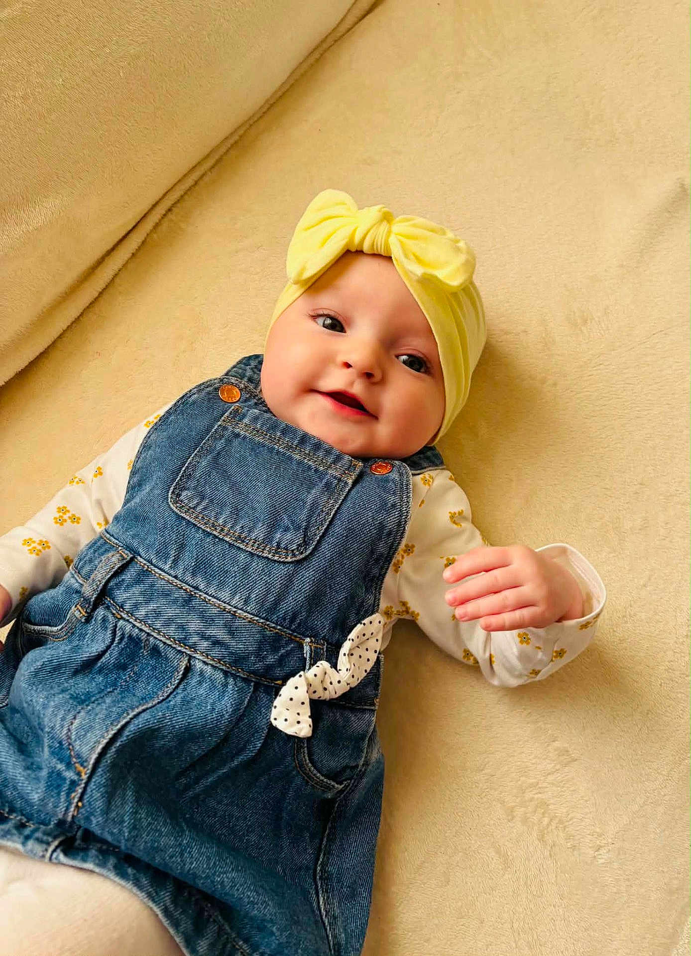 Lyana participe au concours pour gagner de l'argent avec cette photo : baby, infant, child, headband, bow, denim_overalls, onesie, yellow_headband, floral_pattern, blanket, soft_texture, smiling, rosy_cheeks, blue_denim, hand, eyes, cozy, portrait, cute, beige_background