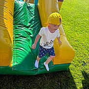 Maël participe au concours pour gagner de l'argent avec cette photo : activity, backward_cap, blue_shorts, casual_clothing, child, fun, grass, green, happy, inflatable_slide, kids, outdoor, play, recreation, slide, summer, sunny_day, white_tshirt, yellow, yellow_cap