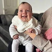 Elodie is registered to the contest to win money with this photo: baby, infant, smiling, happy, person, child, indoor, clothing, pajamas, bib, hand, face, skin, cheeks, sitting, adult, couch, furniture, home, blurred_background