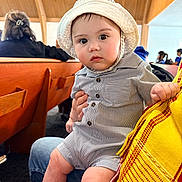 Soren joined the competition — help win amazing prizes! baby, child, hat, striped_clothing, indoor, wooden_bench, person, seated, curious, wide_eyes, lap, hand, people, background, casual, infant, romper, focus, portrait, candid