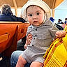 baby, child, hat, striped_clothing, indoor, wooden_bench, person, seated, curious, wide_eyes, lap, hand, people, background, casual, infant, romper, focus, portrait, candid