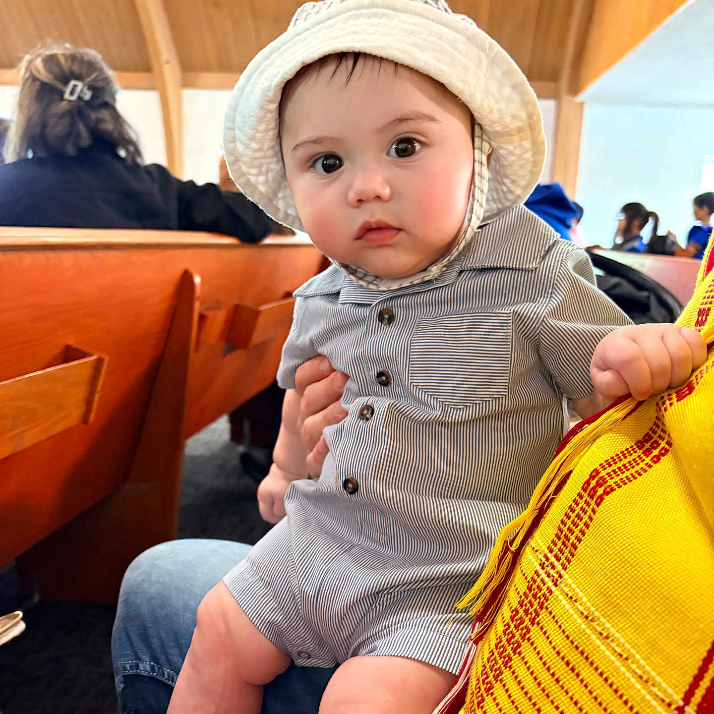 Soren joined the competition — help win amazing prizes! baby, child, hat, striped_clothing, indoor, wooden_bench, person, seated, curious, wide_eyes, lap, hand, people, background, casual, infant, romper, focus, portrait, candid