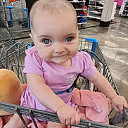 Ellie is registered to the contest to win money with this photo: baby, child, shopping_cart, pink_clothing, blanket, store, retail, clothing_racks, smiling, holding, indoor, floor, person, cute, infant, toddler, face, head, eyes, hands