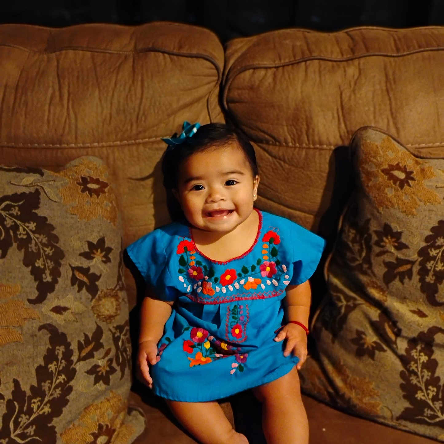 Sophia is registered to the contest to win money with this photo: barefoot, blue_dress, brown_couch, child, couch, cushion, cute, embroidery, face, hair_bow, happy, home, indoor, patterned_cushion, person, portrait, sitting, smile, toddler, young_child