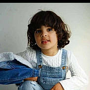Waïl participe au concours pour gagner de l'argent avec cette photo : child, curly_hair, denim, overalls, white_sweater, jacket, portrait, casual_clothing, indoor, light_background, cute, young, person, fashion, smile, face, hand, sitting, studio, curious
