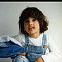 child, curly_hair, denim, overalls, white_sweater, jacket, portrait, casual_clothing, indoor, light_background, cute, young, person, fashion, smile, face, hand, sitting, studio, curious
