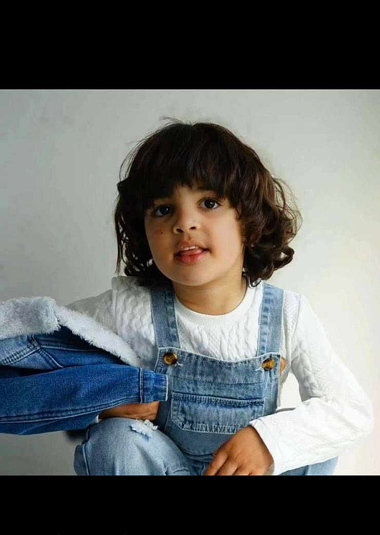 Waïl participe au concours pour gagner de l'argent avec cette photo : child, curly_hair, denim, overalls, white_sweater, jacket, portrait, casual_clothing, indoor, light_background, cute, young, person, fashion, smile, face, hand, sitting, studio, curious