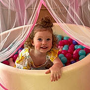 Leni is registered to the contest to win money with this photo: child, toddler, ball_pit, curly_hair, smiling, playful, pink, blue, netting, canopy, indoor, happy, cute, clothing, yellow_dress, person, face, hand, fun, toy