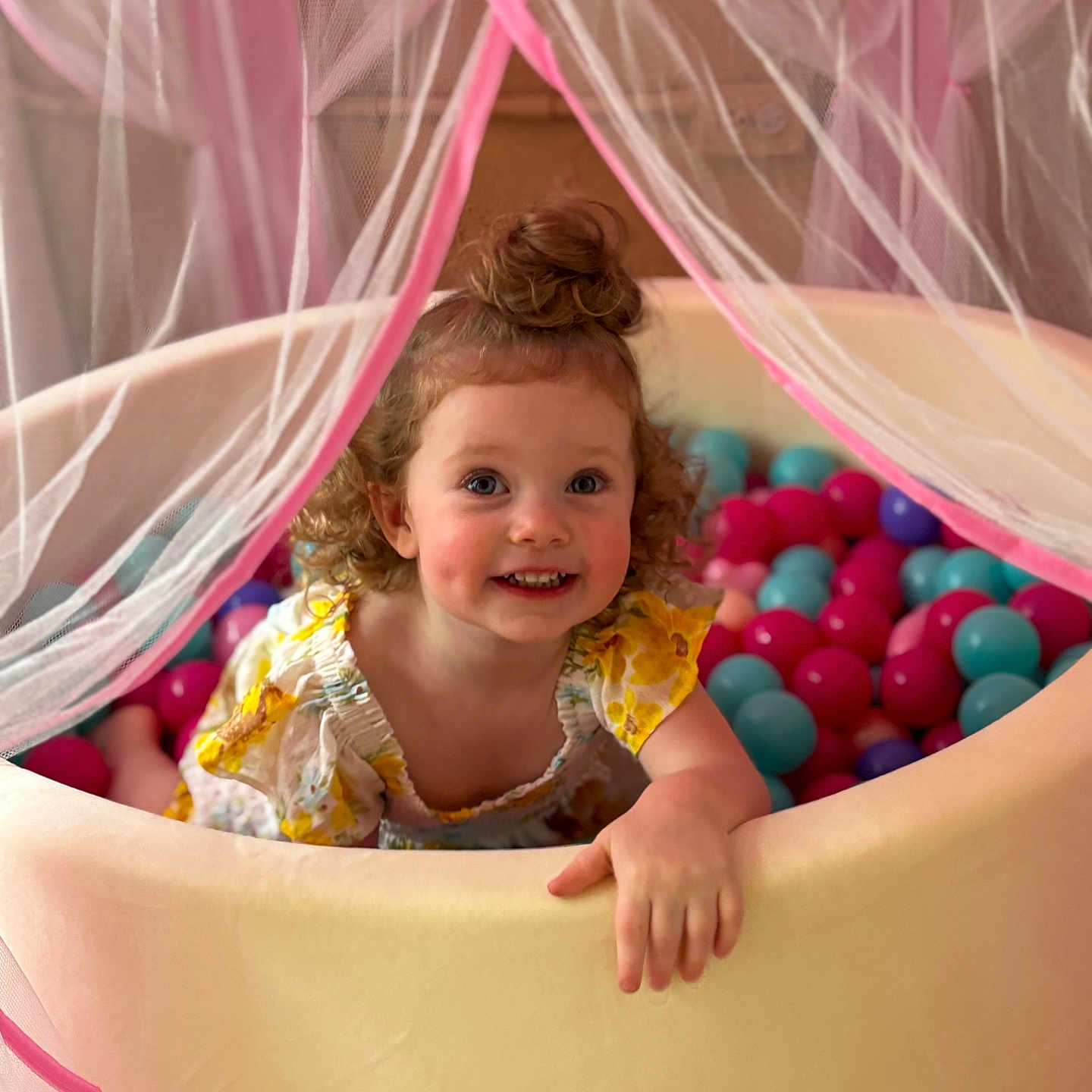 Leni is registered to the contest to win money with this photo: ball_pit, blue, canopy, child, clothing, curly_hair, cute, face, fun, hand, happy, indoor, netting, person, pink, playful, smiling, toddler, toy, yellow_dress