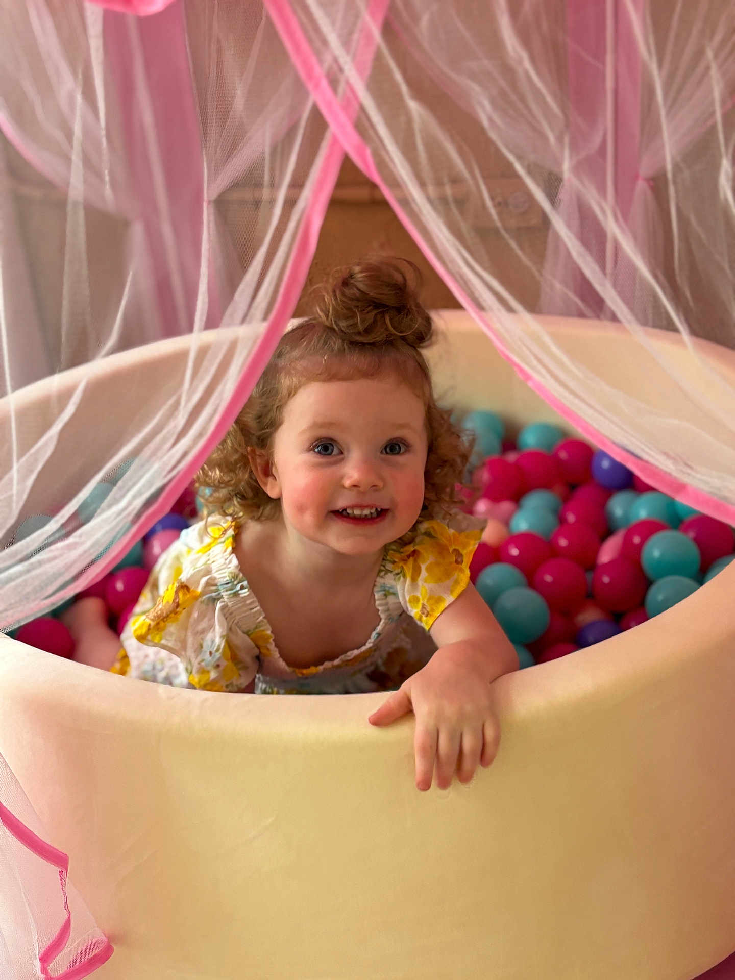 Leni is registered to the contest to win money with this photo: child, toddler, ball_pit, curly_hair, smiling, playful, pink, blue, netting, canopy, indoor, happy, cute, clothing, yellow_dress, person, face, hand, fun, toy