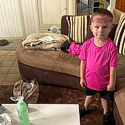 Ayden participe au concours pour gagner de l'argent avec cette photo : bag, black_shorts, blanket, bottle, boy, carpet, casual_clothing, child, couch, expression, floor_tiles, glasses, household_items, indoor, living_room, person, pink_shirt, standing, striped_cushions, table