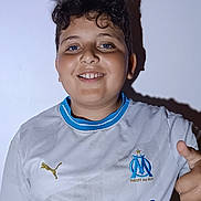 Djessim a rejoint le concours — aidez-le/la à gagner de superbes lots ! child, boy, smile, thumbs_up, curly_hair, white_shirt, sports_jersey, indoor, portrait, happy, blue_eyes, shadow, wall, short_sleeves, casual, person, young, front_view, standing, positive_gesture