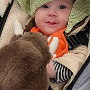 Sabrina is registered to the contest to win money with this photo: baby, child, face, knitted_hat, green_hat, stuffed_animal, buffalo_plush, orange_bib, striped_sleeve, stroller, cozy_clothing, warm_fabric, cute_expression, indoor, portrait, smiling, soft_toys, infant, closeup, winter_wear