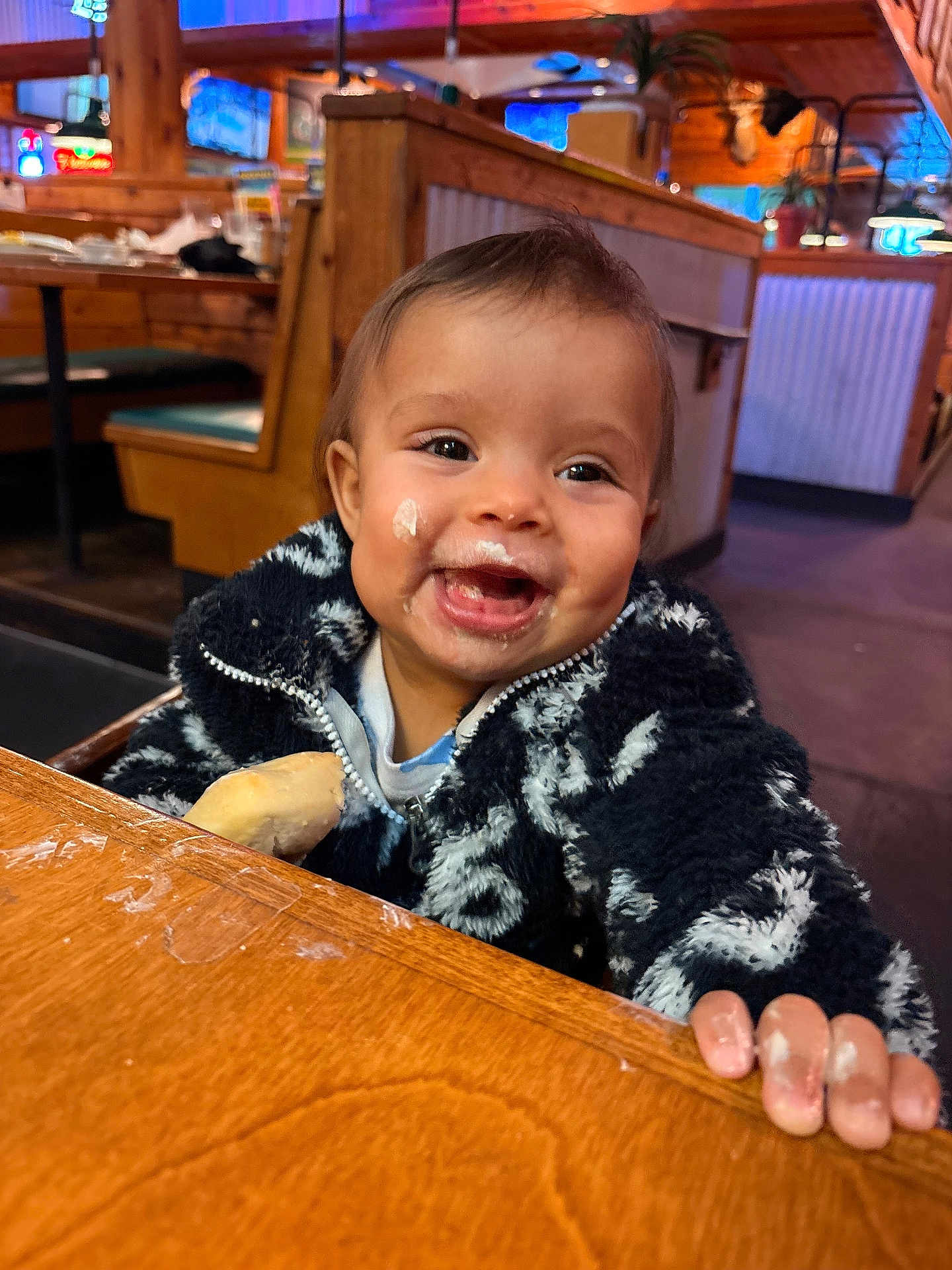 Xiomora is registered to the contest to win money with this photo: child, baby, infant, smiling, messy_face, bread, restaurant, booth, wooden_table, fuzzy_jacket, portrait, dining, happy, hands, cute, candid_photo, food, face, seat, high_emotion