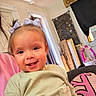 Xiomora is registered to the contest to win money with this photo: toddler, child, smiling, face, hair_bows, green_shirt, indoor, home, couch, pillow, window, curtains, decor, boxes, toy, person, happy, portrait, cute, baby