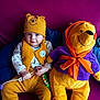 baby, child, plush_toy, winnie_the_pooh, purple_couch, yellow_clothing, hat, vest, cushion, indoor, smiling, person, toy, blue_cushion, orange_scarf, seated, cute, soft_toy, colorful, portrait